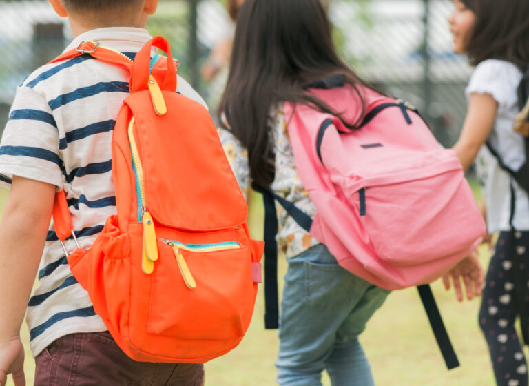 Three pupils of primary school go hand in hand. Boy and girl with school bags behind the back. Beginning of school lessons. Warm day of fall. Back to school. Little first graders.